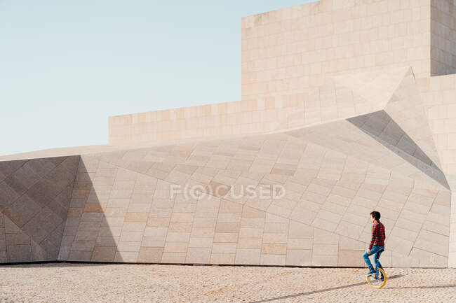 Side view of unrecognizable male riding unicycle against contemporary stone construction of unusual geometric shape — Stock Photo