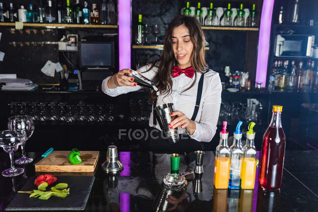 Femme barman en tenue élégante ajoutant des glaçons dans shaker tout en préparant un cocktail debout au comptoir dans le bar moderne — Photo de stock