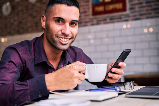Glücklicher hispanischer Mann in violettem Hemd genießt Heißgetränk und surft auf dem Handy in den sozialen Medien, während er während der Arbeit im Café Pause macht — Stockfoto
