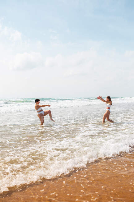 Side view of female friends in swimsuits splashing each other in foamy ocean near sandy beach under blue cloudy sky in sunny day — Stock Photo
