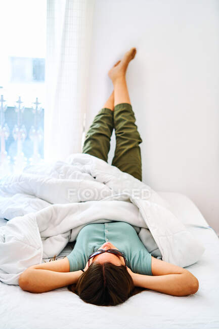 Jeune étudiante en vêtements décontractés et lunettes de soleil allongées avec les jambes levées sur le lit avec couverture et rêvant de vacances d'été — Photo de stock