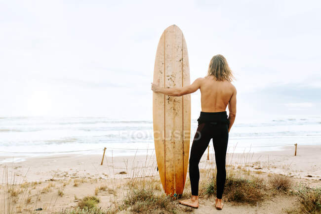 Back view of unrecognizable surfer man with long hair dressed in wetsuit standing with surfboard in sandy dunes — Stock Photo