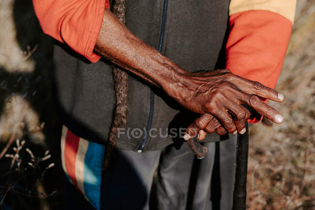 Cultivé vieux rastafari ethnique méconnaissable avec dreadlocks debout avec bâton dans une prairie sèche dans la nature — Photo de stock