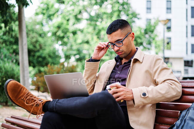 Впевнений молодий іспаномовний чоловік у стильному повсякденному одязі і окулярах відповідає на телефонний дзвінок, сидячи на лавці і працюючи з ноутбуком на вулицях міста. — стокове фото