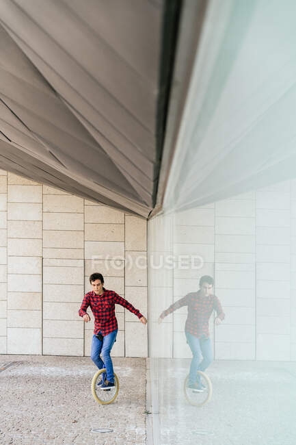 Cuerpo completo de joven hombre activo en camisa a cuadros y jeans realizar truco en monociclo cerca de la pared de vidrio espejo de edificio contemporáneo - foto de stock