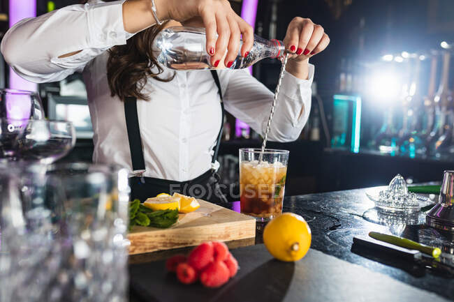 Обрізаний невпізнаваний банкір-жінка в стильному вбранні, що додає рідину з пляшки в склянку і перемішує з довгою ложкою, готуючи коктейль, що стоїть за стійкою в сучасному барі — стокове фото