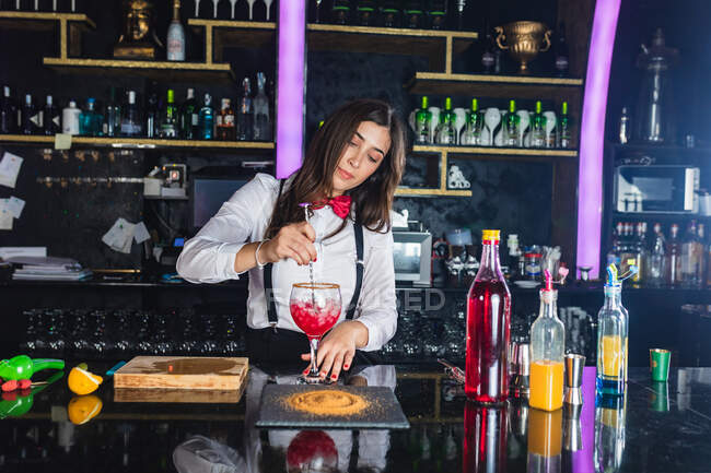Female barkeeper in stylish outfit stirring cocktail in a glass with long spoon standing at counter in modern bar — Stock Photo