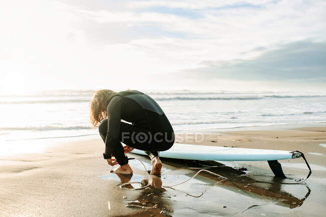Back view of unrecognizable surfer man with long hair dressed in wetsuit  crouched with the surfboard while getting ready to surf on the beach during  sunrise in the background — Men, seacoast -