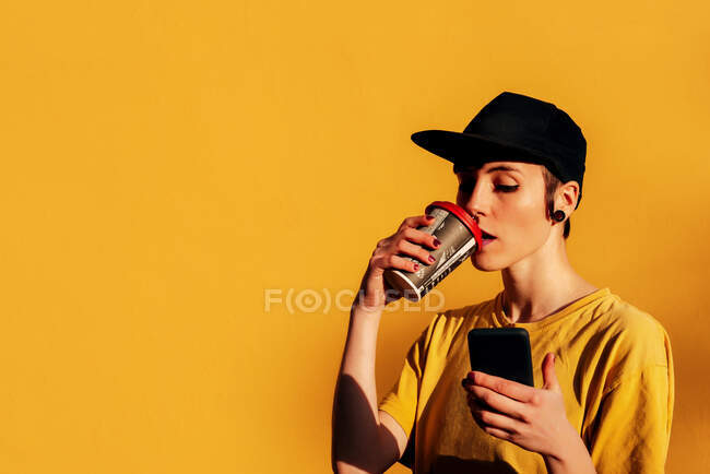 Jovem mulher informal na moda tampão bebendo takeaway bebida quente e navegação smartphone perto de fundo amarelo — Fotografia de Stock