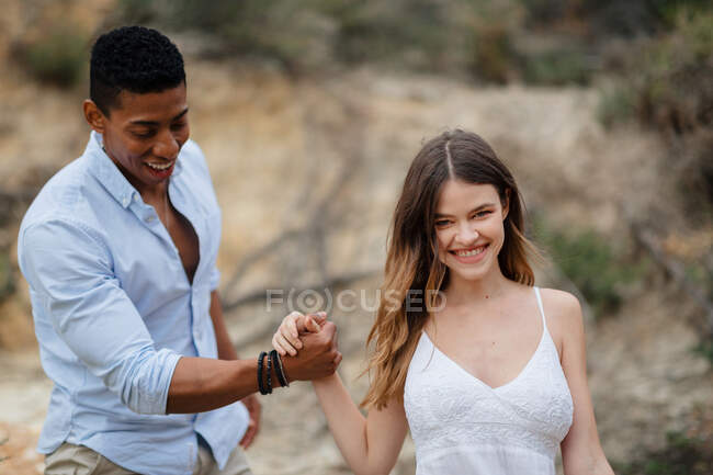 Delighted multiracial bridge and groom holding hands and walking along sandy hill on wedding day in nature — Stock Photo