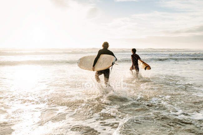Обратный вид неузнаваемых друзей-серферов, одетых в гидрокостюмы, идущих с досками для серфинга к воде, чтобы поймать волну на пляже во время восхода солнца — стоковое фото