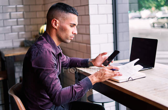 Vista lateral del joven empresario hispano guapo comprobando la información en el teléfono inteligente y escribiendo notas en el planificador mientras trabaja en la mesa con el ordenador portátil - foto de stock