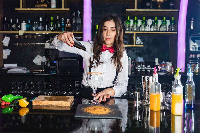 Female barkeeper in stylish outfit adding ice cubes into a glass while preparing cocktail standing at counter in modern bar — Stock Photo