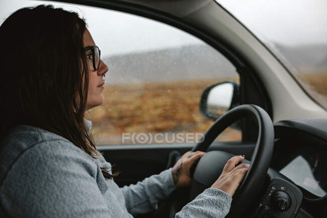 Vue latérale de la jeune femme en lunettes avec une automobile de conduite perçant entre les terres sauvages avec des collines par temps pluvieux — Photo de stock