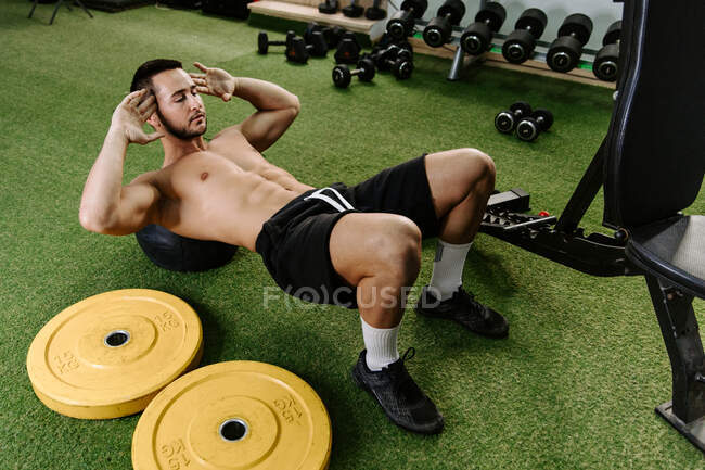 Hombre fuerte enfocado con torso desnudo haciendo abdominales crujidos mientras entrena en el gimnasio - foto de stock