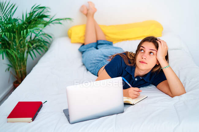 Молодая студентка лежит на кровати и пишет в блокноте во время обучения онлайн с ноутбуком во время дистанционного обучения дома — стоковое фото