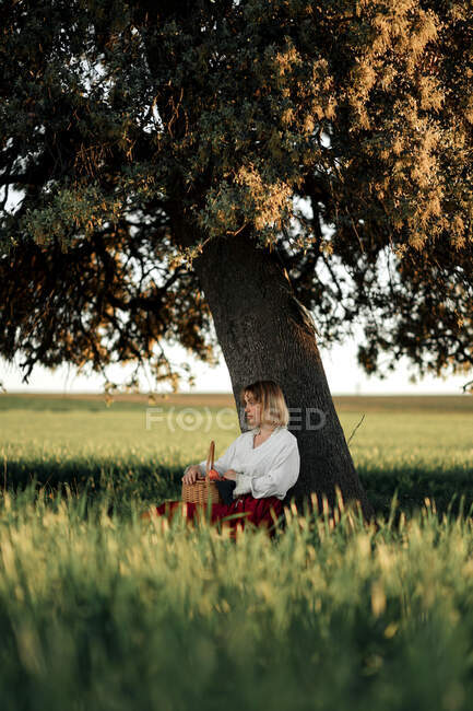 Young female in old fashioned white blouse and skirt holding wicker basket full of fresh apples while resting near tree in summer day in countryside — Stock Photo