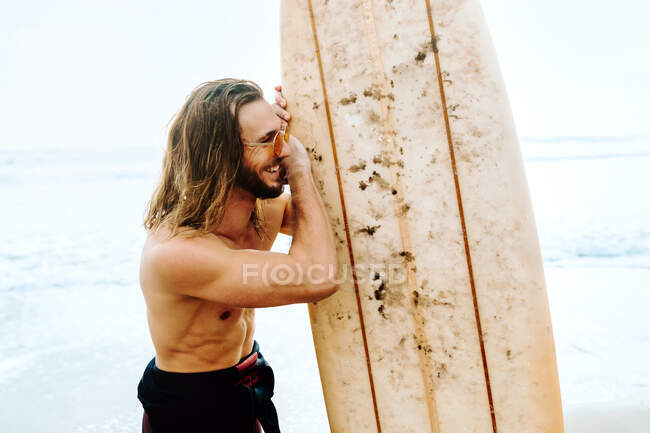 Young cheerful handsome surfer man with long hair dressed in wetsuit and stylish sunglasses standing looking away with surfboard towards the water to catch a wave on the beach — Stock Photo