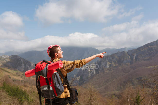 Back view of delighted female traveler with backpack standing in mountains and pointing away — Stock Photo