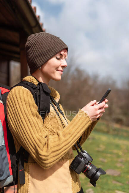 Вид збоку весела жінка-дослідник з рюкзаком, що стоїть на природі біля будинку та переглядає мобільний телефон — стокове фото