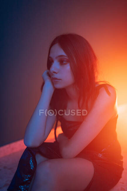 Спокойная молодая модель в платье сидит на полу и опирается на руку, глядя в темную студию с красочными огнями — стоковое фото