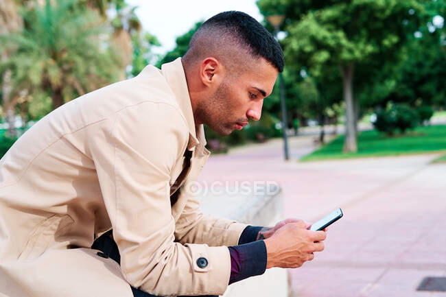 Низький кут позитивно одягненого молодого іспанського бізнесмена СМС на смартфоні і обговорення новин на міській вулиці з сучасними будівлями на задньому плані — стокове фото