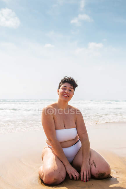 Sorridente giovane plus size femminile in costume da bagno seduto sulla spiaggia sabbiosa guardando la fotocamera vicino all'oceano schiumoso sotto il cielo nuvoloso blu alla luce del giorno — Foto stock