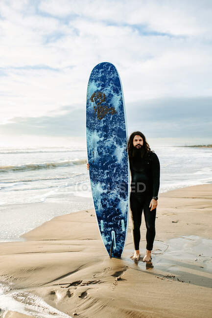 Surfer man with long hair and beard dressed in wetsuit standing looking at camera with the surfboard on the beach during sunrise in the background — Stock Photo