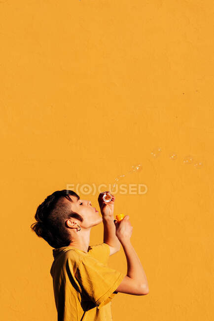 Femme moderne avec des bulles de savon perçant soufflant avec les yeux fermés sur la journée ensoleillée contre le mur jaune — Photo de stock