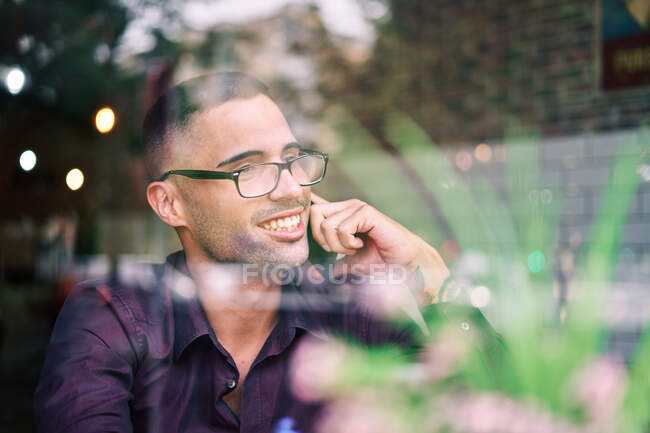 Empresario hispano inteligente en gafas tomando notas en bloc de notas y contestando llamadas telefónicas mientras está sentado detrás de la ventana en la cafetería - foto de stock