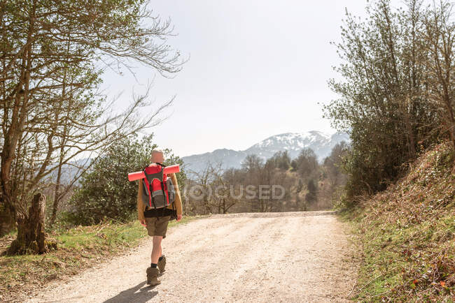 Visão traseira do explorador com mochila andando na estrada arenosa que leva para terras altas no dia ensolarado — Fotografia de Stock