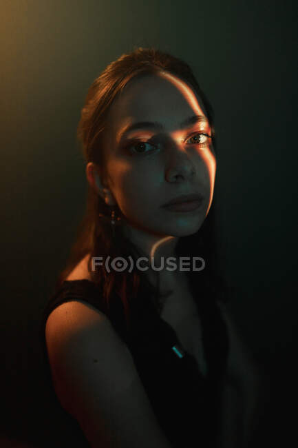 Joven modelo femenino pensativo con proyección de luz en la cara sentado en estudio oscuro y mirando hacia otro lado - foto de stock