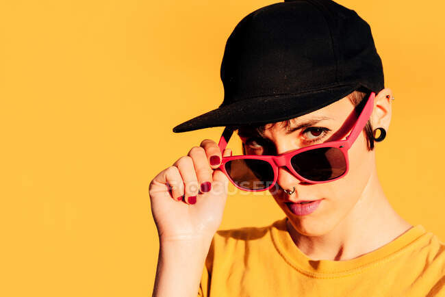 Mulher confiante em boné preto olhando para a câmera e ajustando óculos de sol rosa vívidos contra fundo amarelo — Fotografia de Stock