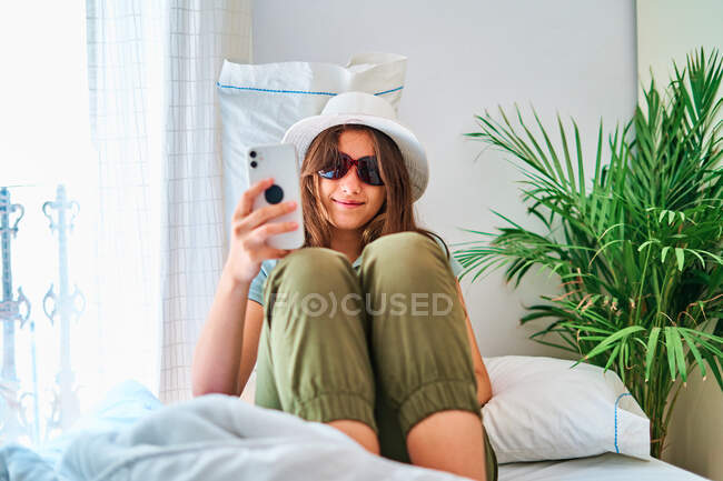 Lächelnde junge Frau in Sommerkleidung und Hut mit Sonnenbrille sitzt auf dem Bett und macht ein Selfie mit dem Smartphone, während sie Zeit allein zu Hause verbringt — Stockfoto