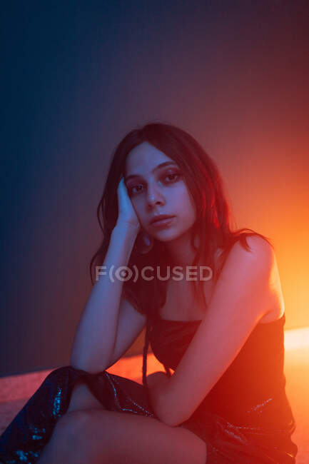 Tranquil modelo femenina joven en vestido sentado en el suelo y apoyado en la mano mientras mira a la cámara en el estudio oscuro con luces de colores - foto de stock