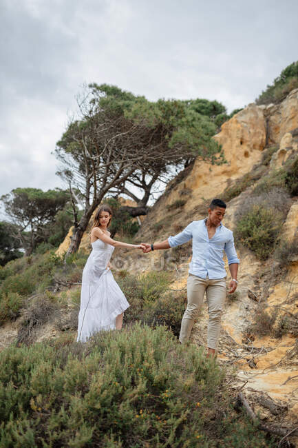 Entzückte multiethnische Brücke und Bräutigam Händchen haltend und entlang sandiger Hügel am Hochzeitstag in der Natur — Stockfoto