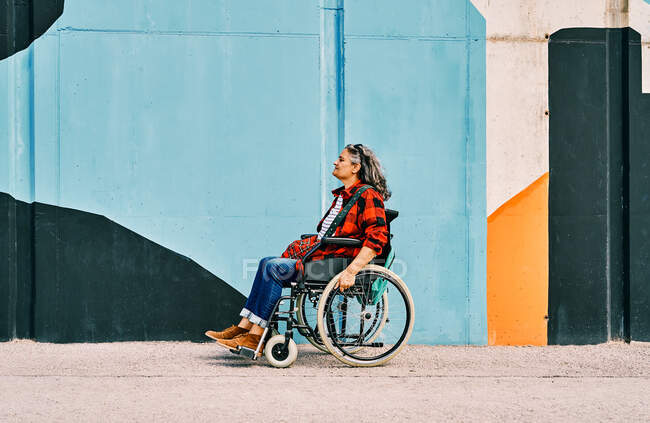 Vista lateral de cuerpo completo de la silla de ruedas femenina madura discapacitada independiente cerca de la pared pintada en la calle - foto de stock