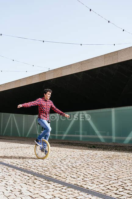 Полный вид на тело активного молодого мужчины в клетчатой рубашке и джинсах, выполняющего трюк на одноколесном велосипеде возле зеркальной стеклянной стены современного здания — стоковое фото