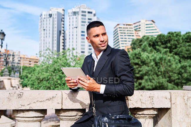 Vista lateral del joven gerente ejecutivo hispano enfocado en traje elegante con información de lectura de bolsos en la tableta mientras está parado cerca de la barandilla en la terraza urbana - foto de stock