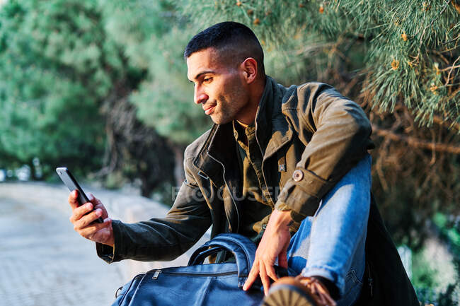 Hispânico cara em roupas casuais olhando para longe e mensagens de texto no telefone enquanto estava perto de barreira e árvore conífera no parque — Fotografia de Stock