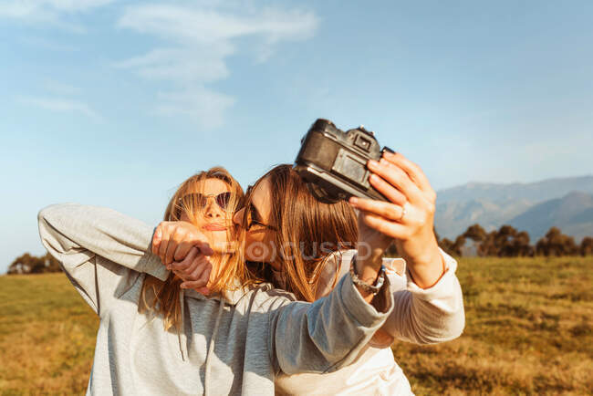 Alegre jóvenes novias en gafas de sol tomando auto foto con cámara de película analógica y besándose en el campo de las montañas - foto de stock