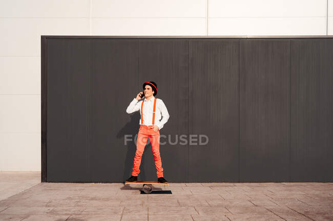 Voller Körper von fröhlichen jungen männlichen Zirkusartisten, die auf dem Balancierbrett an einer grauen Wand stehen und mit dem Smartphone sprechen — Stockfoto