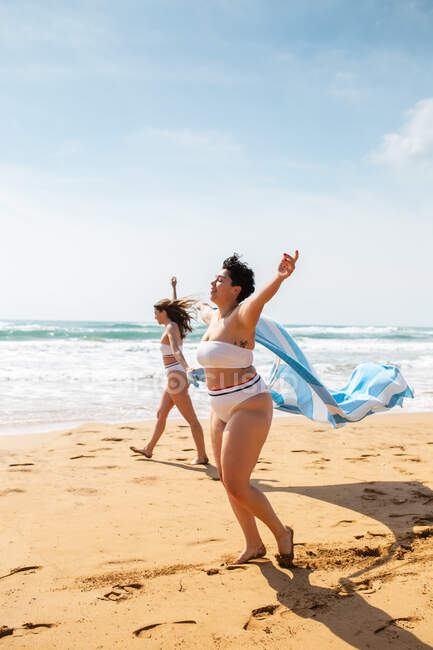 Visão lateral de corpo inteiro de amigas em trajes de banho passeando na costa arenosa com toalha perto do oceano sob o céu azul nublado em dia ensolarado — Fotografia de Stock