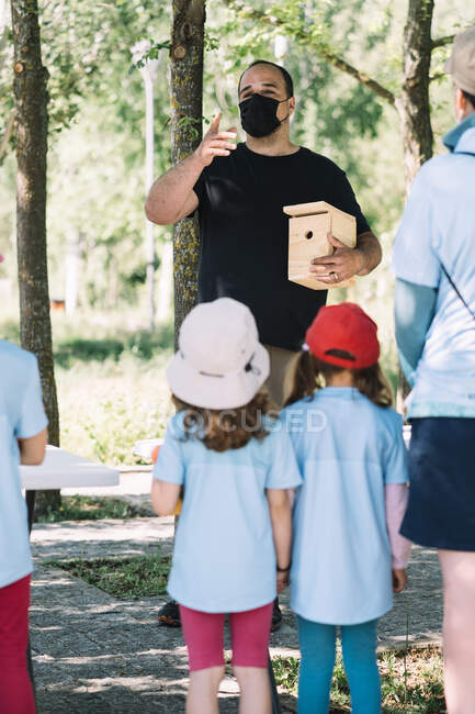 Человек в защитной маске с деревянной коробкой для гнездования ручной работы, общающийся с группой детей-добровольцев, собирающихся вместе в летнем парке — стоковое фото