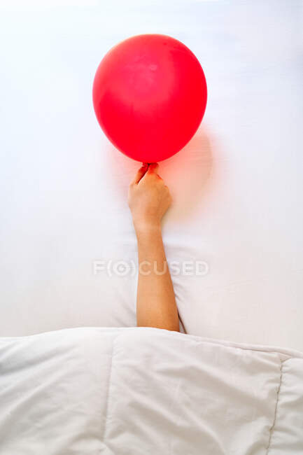 Cropped unrecognizable tired person with red balloon in hand sleeping in bed with white sheets after party — Stock Photo