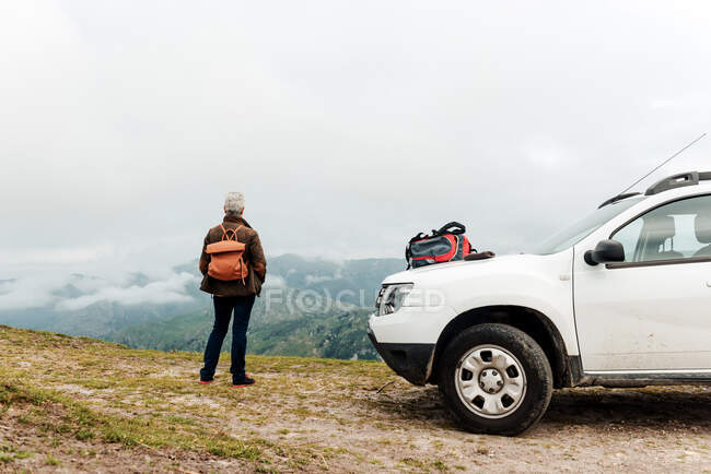 Visão traseira da mulher idosa anônima com mochila de pé perto do veículo branco e admirando a montanha durante a viagem de carro em terras altas no dia cinza nublado — Fotografia de Stock