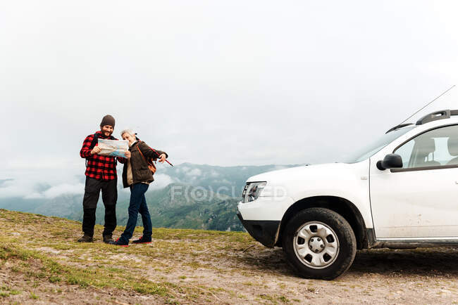 Senior woman and adult man examining map together while standing near white vehicle during road trip in mountains — Stock Photo