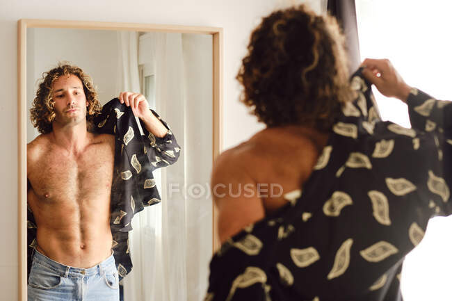 Handsome male with strong torso putting on shirt in front of mirror in room at home — Stock Photo