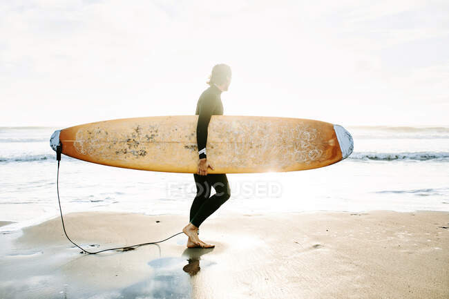 Vista lateral del surfista vestido con traje de neopreno caminando por la playa con la tabla de surf en la mañana con salida del sol en el fondo - foto de stock