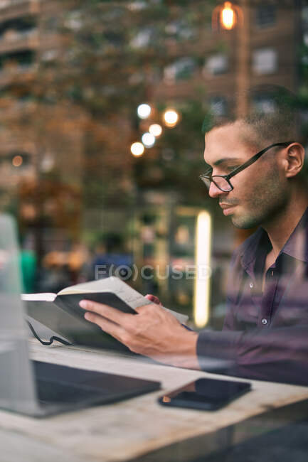 Homem hispânico inteligente em óculos ler notas no planejador enquanto sentado à mesa atrás da janela e trabalhando em projeto de negócios na cafetaria — Fotografia de Stock